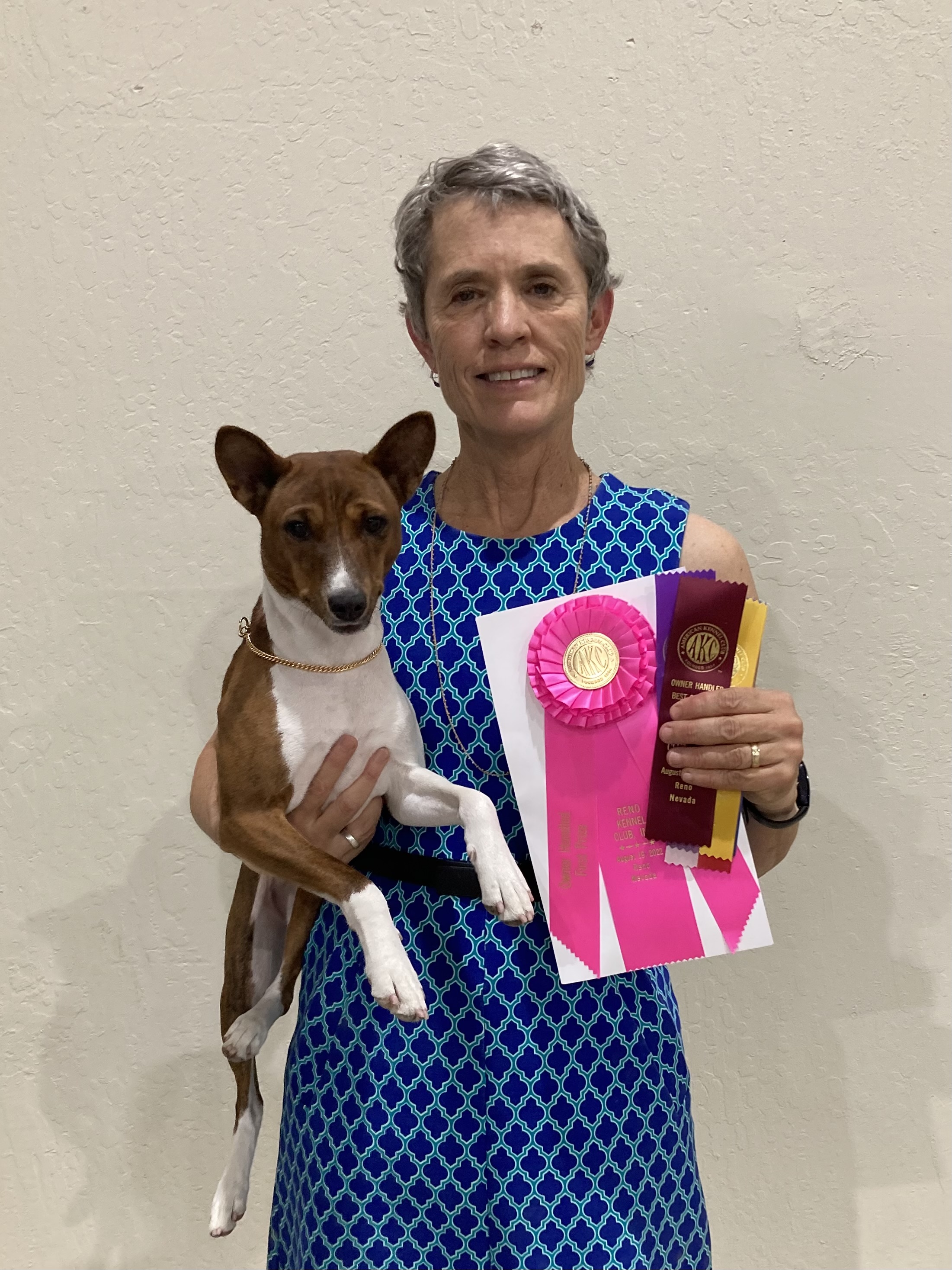 A older women holding her basenji dog in one arm while holding out the awards it won with the other hand